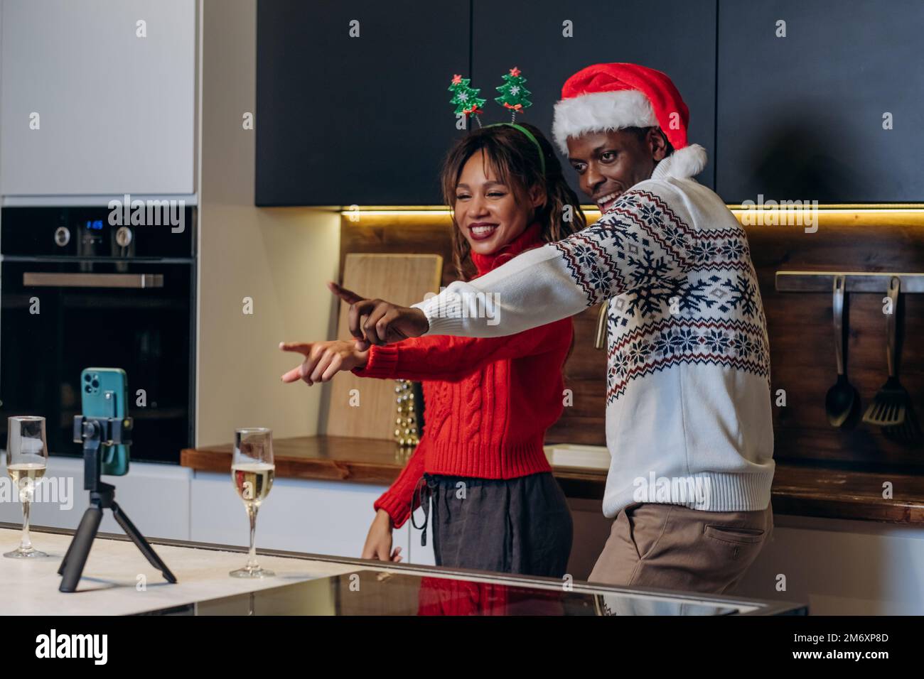 Ein afroamerikanischer Mann in Santa hat filmt ein Weihnachtsvideo mit einer lockigen Freundin im Strickpullover, die in der Küche in festlicher Atmosphäre steht Stockfoto