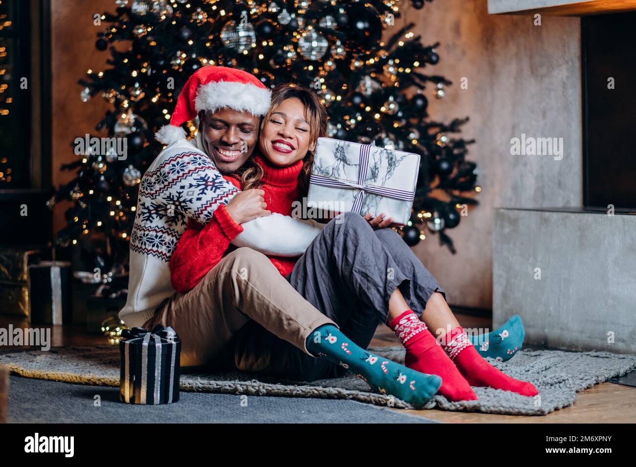Eine glückliche Frau und ein schwarzer Mann in festlichen Pullover, die sich umarmen, Geschenke halten und auf dem Teppich neben dem Weihnachtsbaum mit Ornamenten und Girlande sitzen Stockfoto
