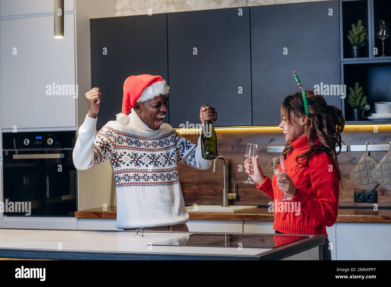 Eine junge Frau und ein afroamerikanischer Mann, die festliche Hüte und Strickpullover tragen, trinken Champagner aus Gläsern, die Weihnachten in der Küche feiern Stockfoto