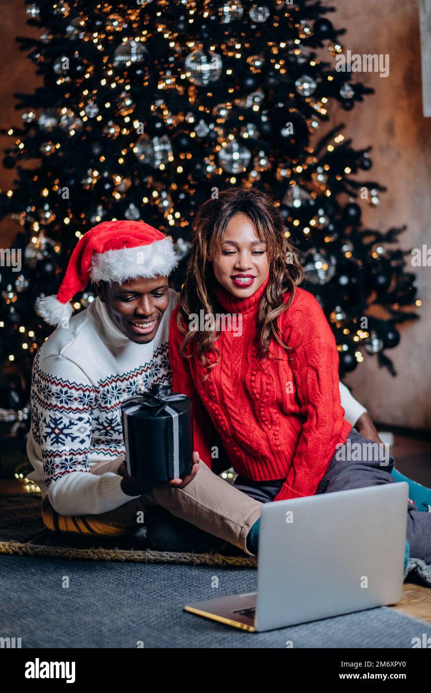 Schwarzer Mann und weiße Frau in festlichen Pullover unterhalten sich über einen Laptop und zeigen die Geschenkbox mit Verzierungen vor dem Weihnachtsbaum Stockfoto