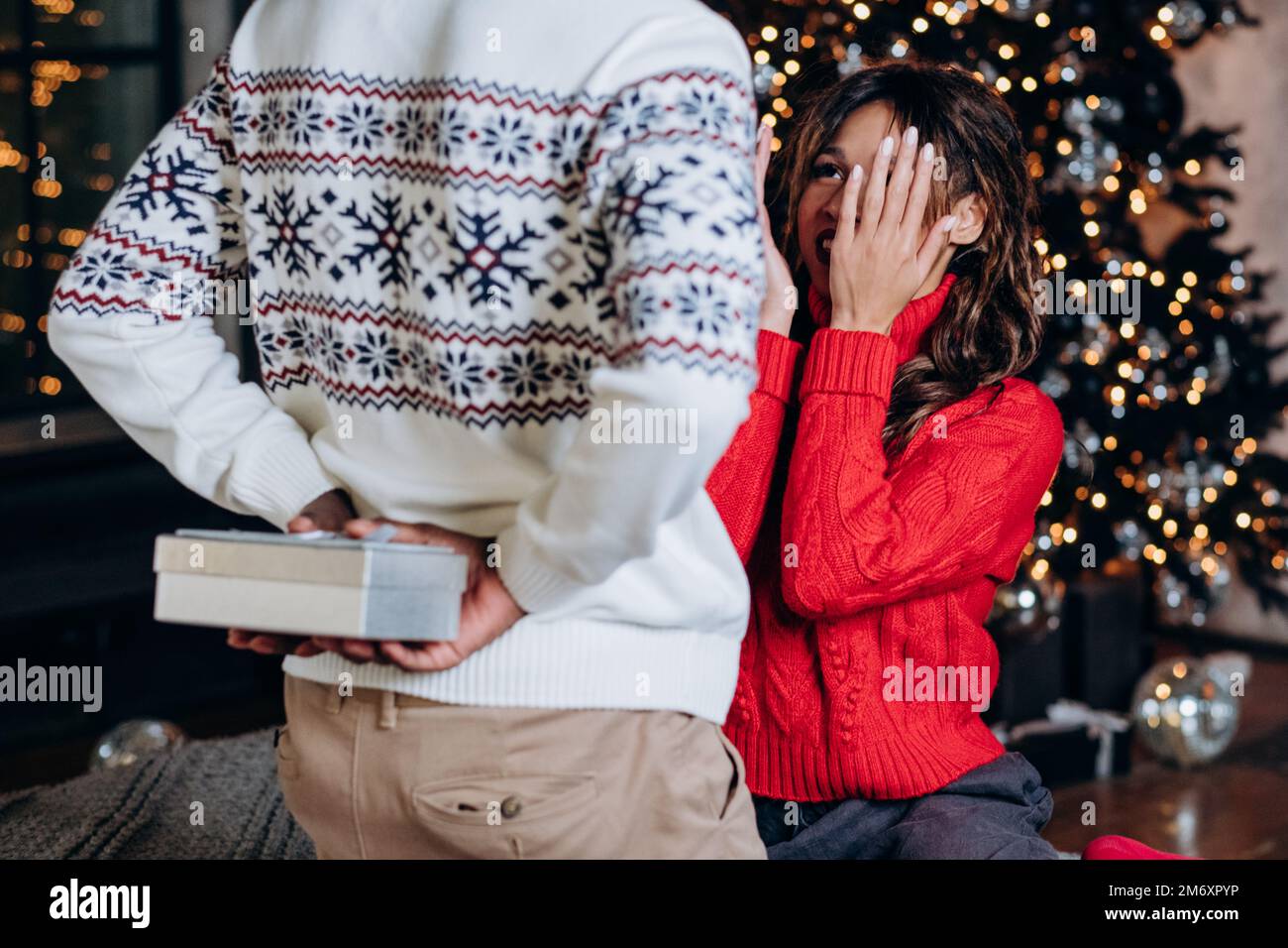 Ein afroamerikanischer Mann in einem warmen bedruckten Pullover überrascht eine lockige brünette Frau mit einer Geschenkbox hinter dem Rücken gegen den Weihnachtsbaum Stockfoto