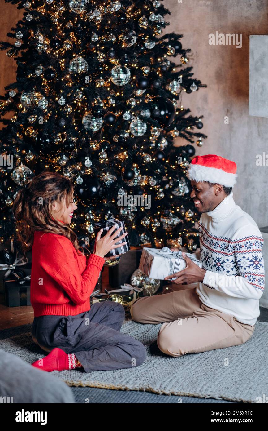 Ein junges glückliches Paar schenkt einander ein Lächeln und sitzt auf einem gestrickten Teppich neben einem Weihnachtsbaum, der mit Ornamenten und Girlande dekoriert ist Stockfoto