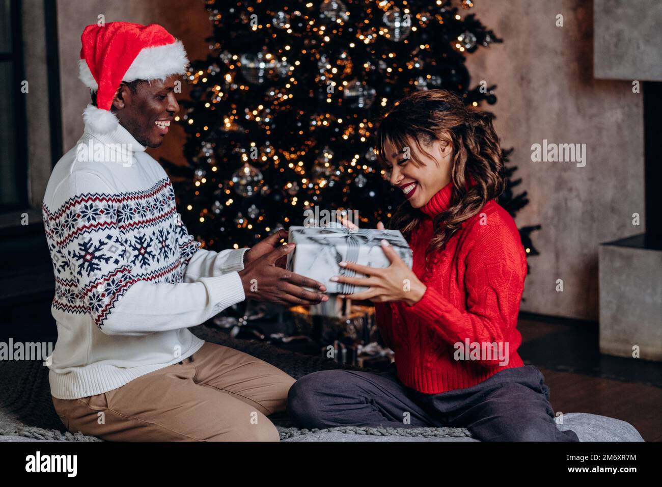 Ein afroamerikanischer Mann mit Weihnachtsmann-Hut überrascht eine langhaarige Freundin mit einem Geschenk, das auf dem Boden neben einem dekorierten Weihnachtsbaum sitzt Stockfoto
