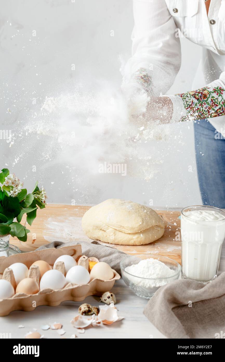 Weibliche Hände klatschen und Mehlspritzer über dem Teig auf hellem Hintergrund machen. Eier und Zutaten sind im Vordergrund. Leerer Bereich für Text Stockfoto