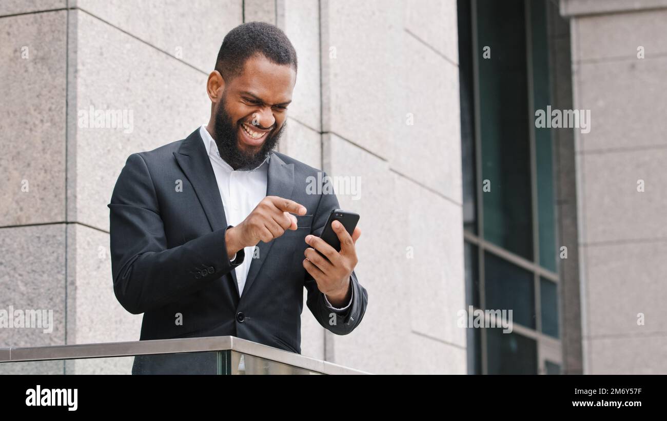 Männlicher, ethnisch erfolgreicher, lächelnder, bärtiger afroamerikanischer Unternehmer, Unternehmer, der mit dem Sieg des Mobiltelefons zufrieden ist, mit dem gute Nachrichten gewinnen Stockfoto