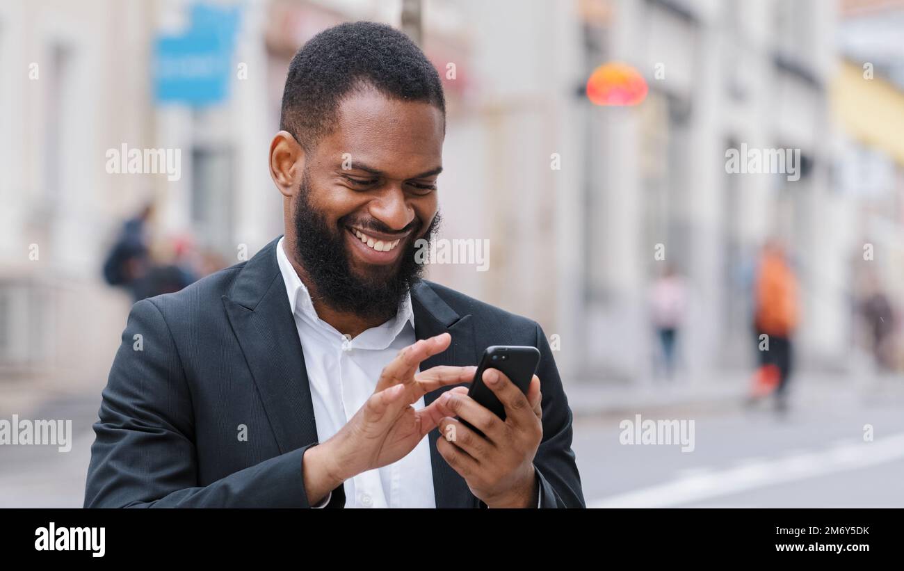 Lächelnder, afroamerikanischer, glücklicher, bärtiger Geschäftsmann, Geschäftsführer mit Smartphone in der Stadt, chattet mit SMS und liest geschäftliche Chats Stockfoto