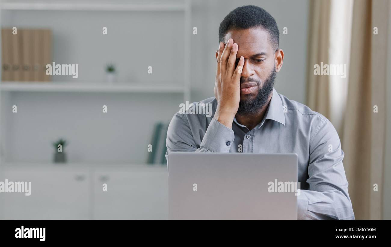 Gestresster kranker afroamerikanischer, bärtiger Mann leidet unter Kopfschmerzen, Schmerzen, Büroangestellter, Geschäftsmann mit Laptop, traurig verärgert, frustriert über Stockfoto