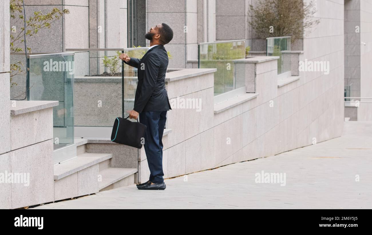 Ein afroamerikanischer ethnischer Kandidat bietet ein Arbeitsgespräch an, der neben dem Bürogebäude steht und auf die Armbanduhr schaut. Geschäftsmann-Unternehmer, beeilen Sie sich Stockfoto