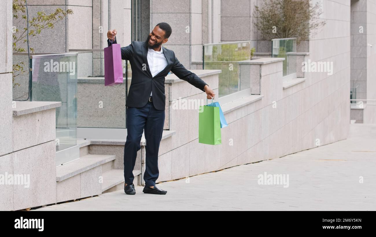 Millennial männliche Kunden, die aus ethnischen Gründen Käuferinnen sind, Geschäftsmann aus Afroamerikanern, der aus dem Einkaufszentrum geht, einen Boutique-Spaziergang in der Stadt macht, mit glücklichen Einkaufstaschen Stockfoto
