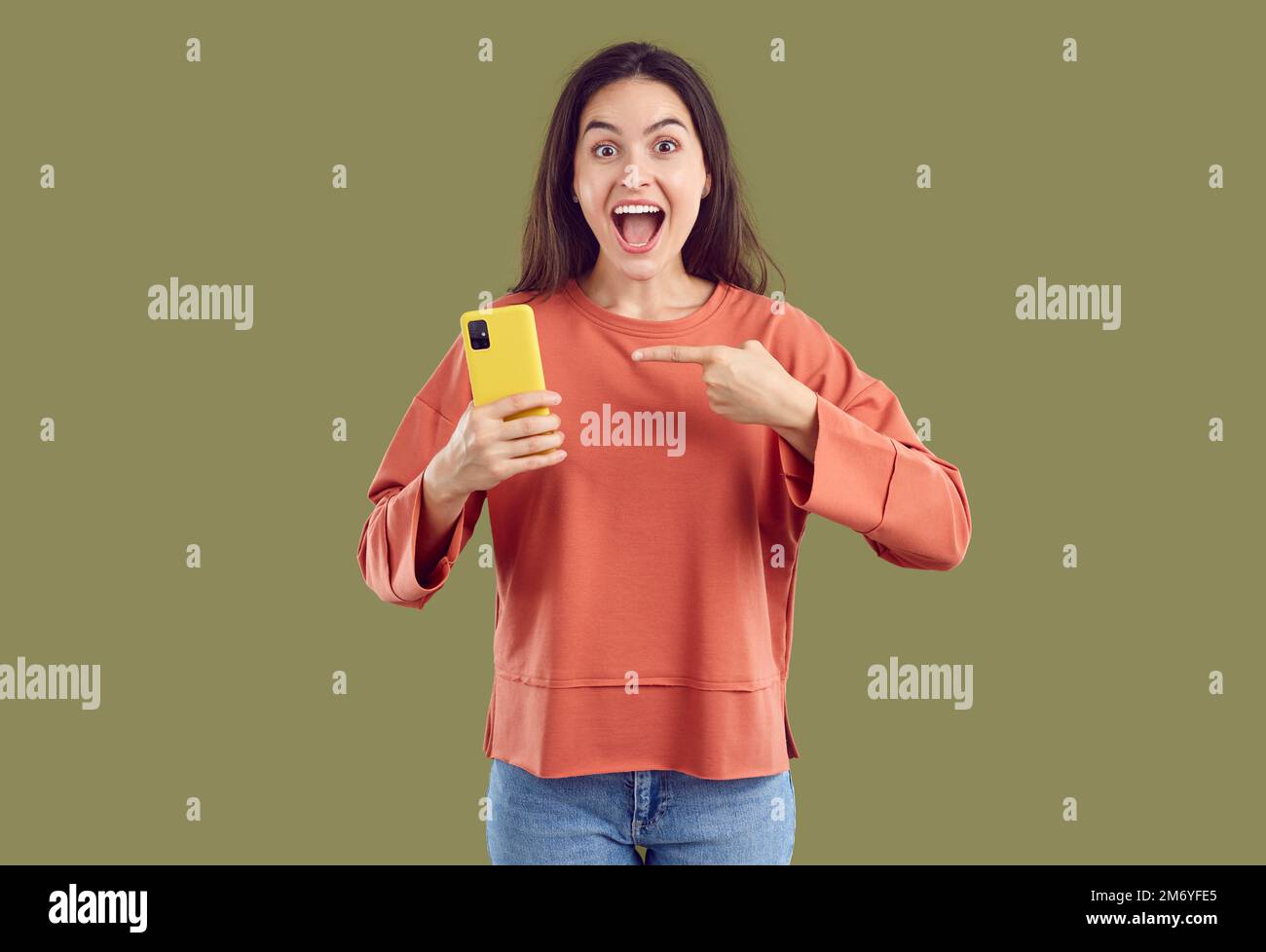 Eine verblüffte junge brünette Frau in lässiger Kleidung zeigt auf das Smartphone im Khaki-Hintergrund. Stockfoto