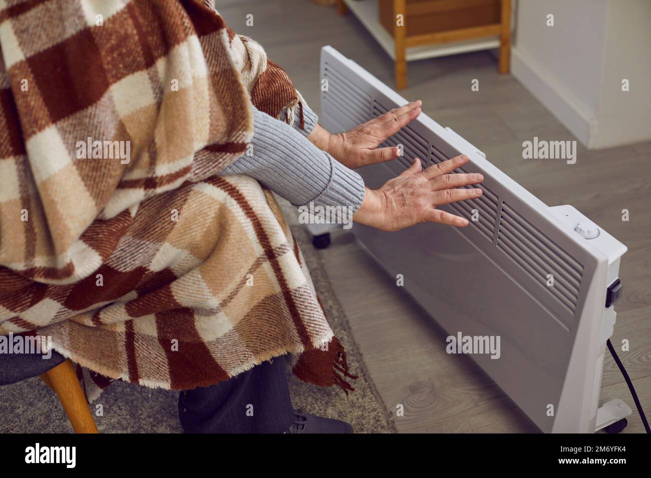 Ein älterer Mann zu Hause erwärmt sich die Hände an der elektrischen Heizung aufgrund von Problemen mit der Zentralheizung. Stockfoto