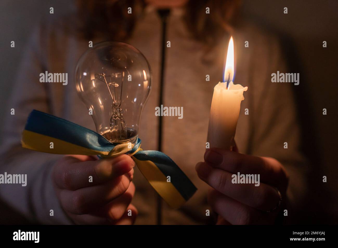 Stromausfall. Stromausfallkonzept. Mädchen hält eine brennende Kerze und eine elektrische Glühbirne mit einem gelb-blauen Band (nationales Symbol der Ukraine) Stockfoto