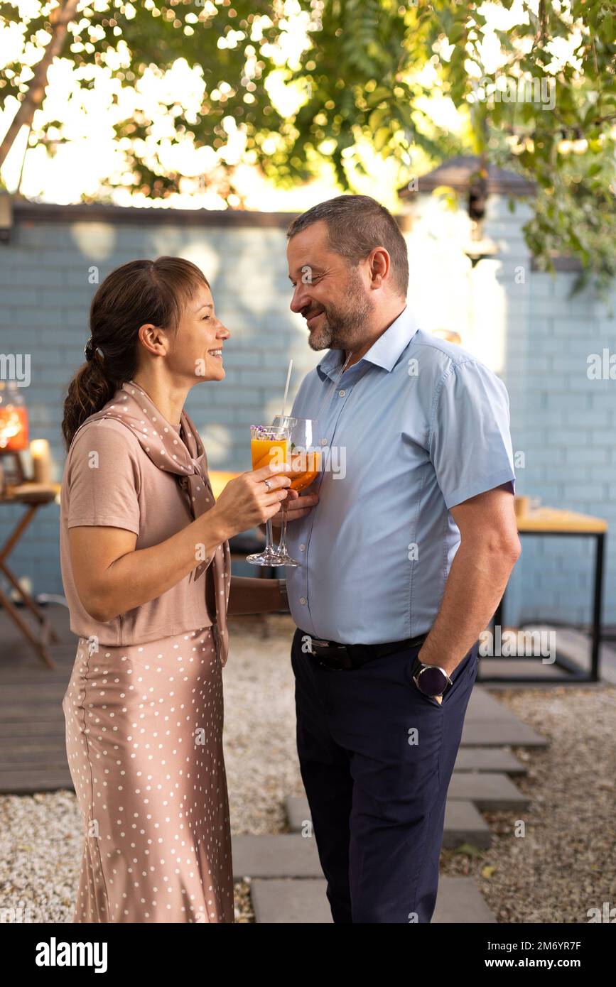 Ein glückliches Paar, das sich im Terrassengarten unterhalten und geflirtet hat und Cocktails serviert. Menschen, die bellini-Cocktails und Aperol Spritz zusammen in Outdoor s anstoßen Stockfoto