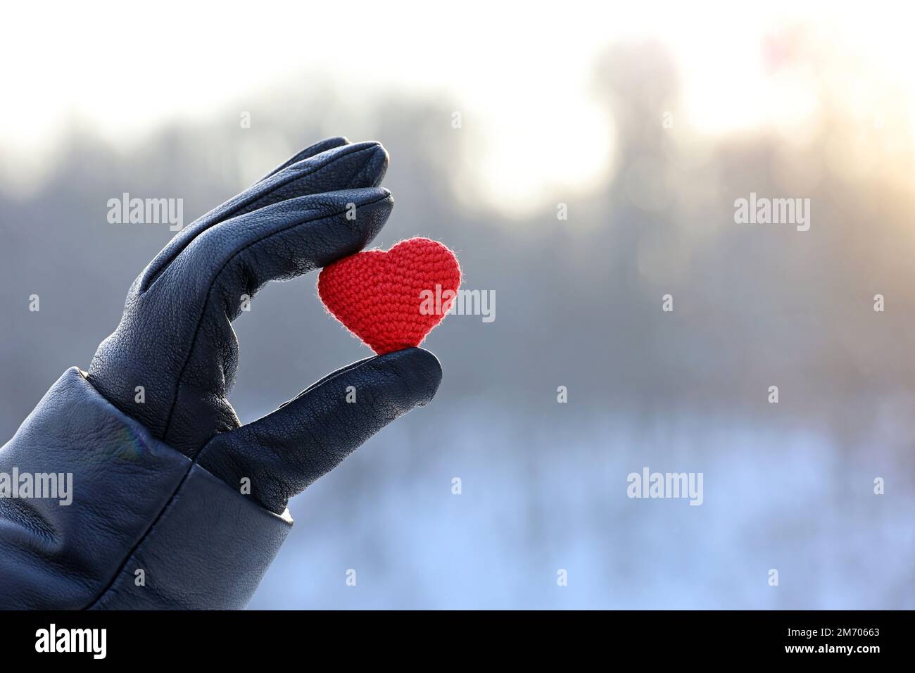 Rotes gestricktes Herz in weiblicher Hand in schwarzem Lederhandschuh auf Schnee- und Sonnenhintergrund. Konzept der romantischen Liebe, Valentinstag, Winterwetter Stockfoto