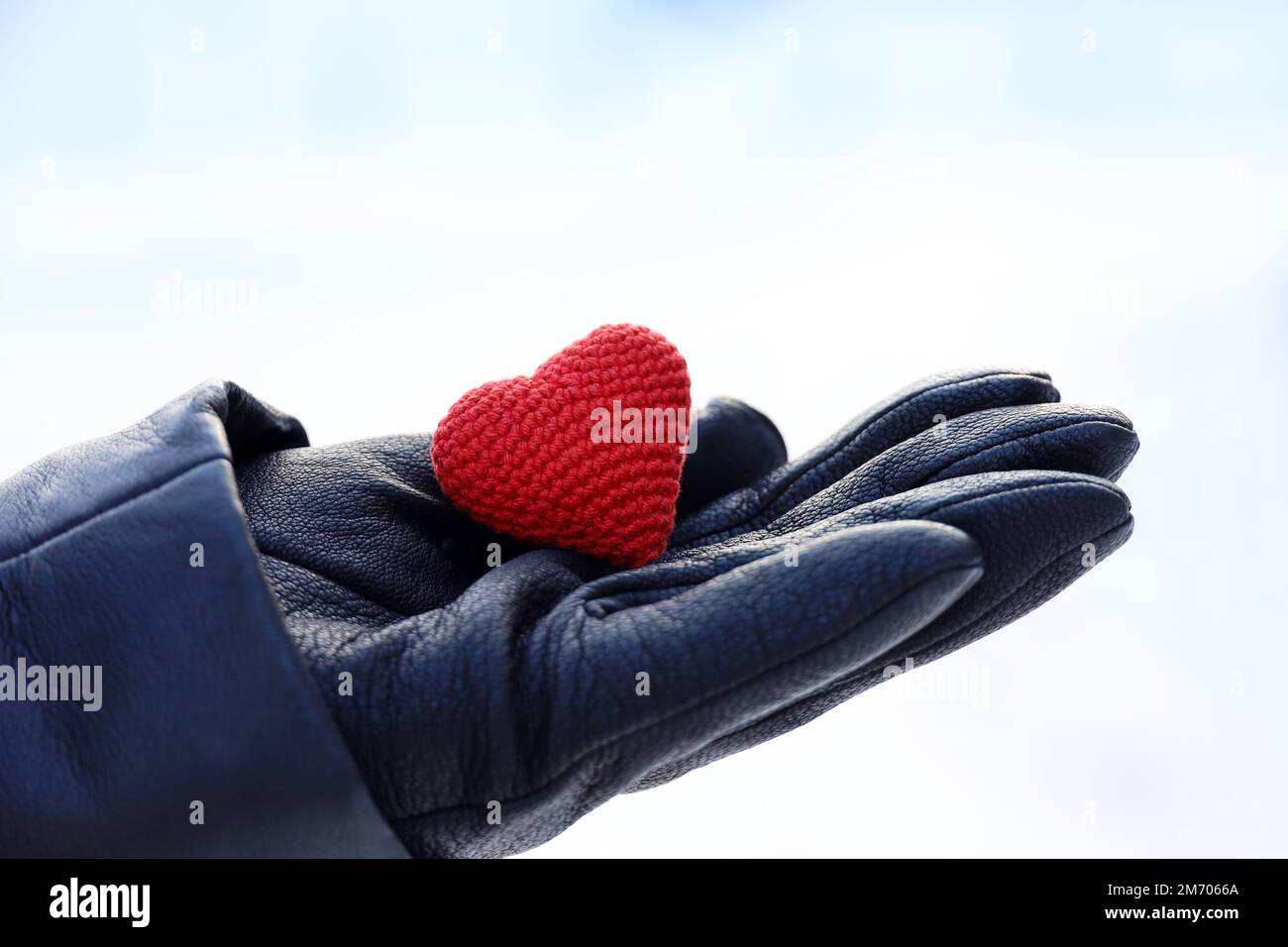 Rotes Strickherz in weiblicher Handfläche in schwarzem Lederhandschuh auf Schneehintergrund. Konzept der romantischen Liebe, Valentinstag, Winterwetter Stockfoto