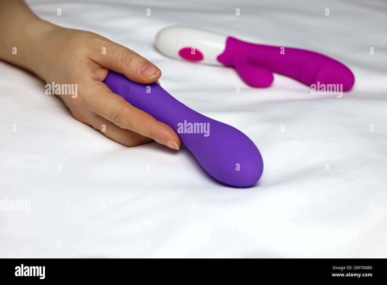 Sexspielzeug in weiblicher Hand auf weißem Laken. Eine Frau, die auf einem  Bett liegt, mit einem lilafarbenen Silikon-Dildo und einem roten Vibrator  Stockfotografie