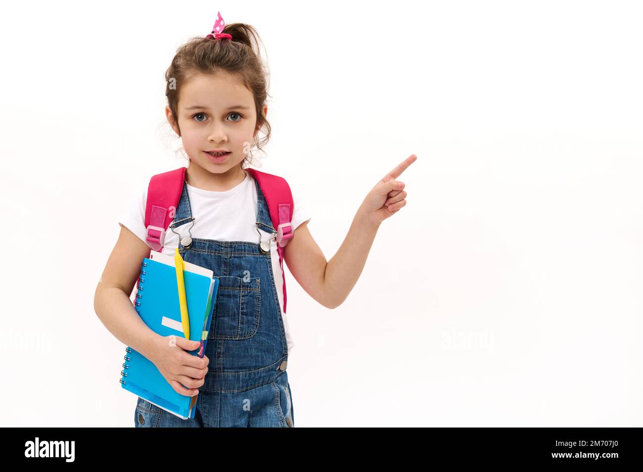 Grundschülerin, hübsches Mädchen mit Copybook und Rucksackspitzen Zeigefinger auf dem Kopierfeld auf Weiß. Zurück in die Schule Stockfoto