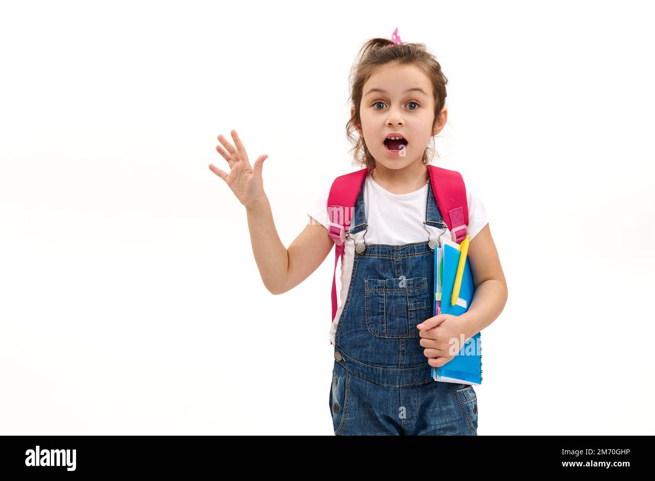 Ein überraschtes kleines Mädchen, eine Grundschülerin, die Überraschung ausdrückt, mit Schulutensilien vor weißem Hintergrund posiert Stockfoto