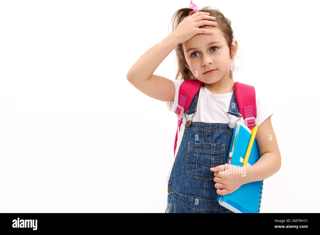 Grundschüler, niedliches kleines Mädchen, das Hand auf die Stirn legt, mit pinkfarbenem Rucksack und Copybook auf Weiß posiert Stockfoto