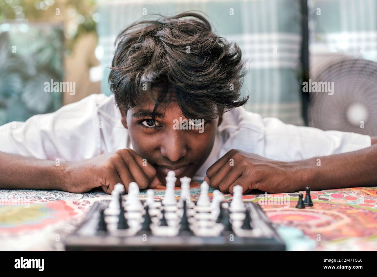 Ein Junge indischer Herkunft starrt in die Kamera, während er Schach spielt. Vorderansicht. Porträt eines indischen Jungen mit Schachbrett. Stockfoto