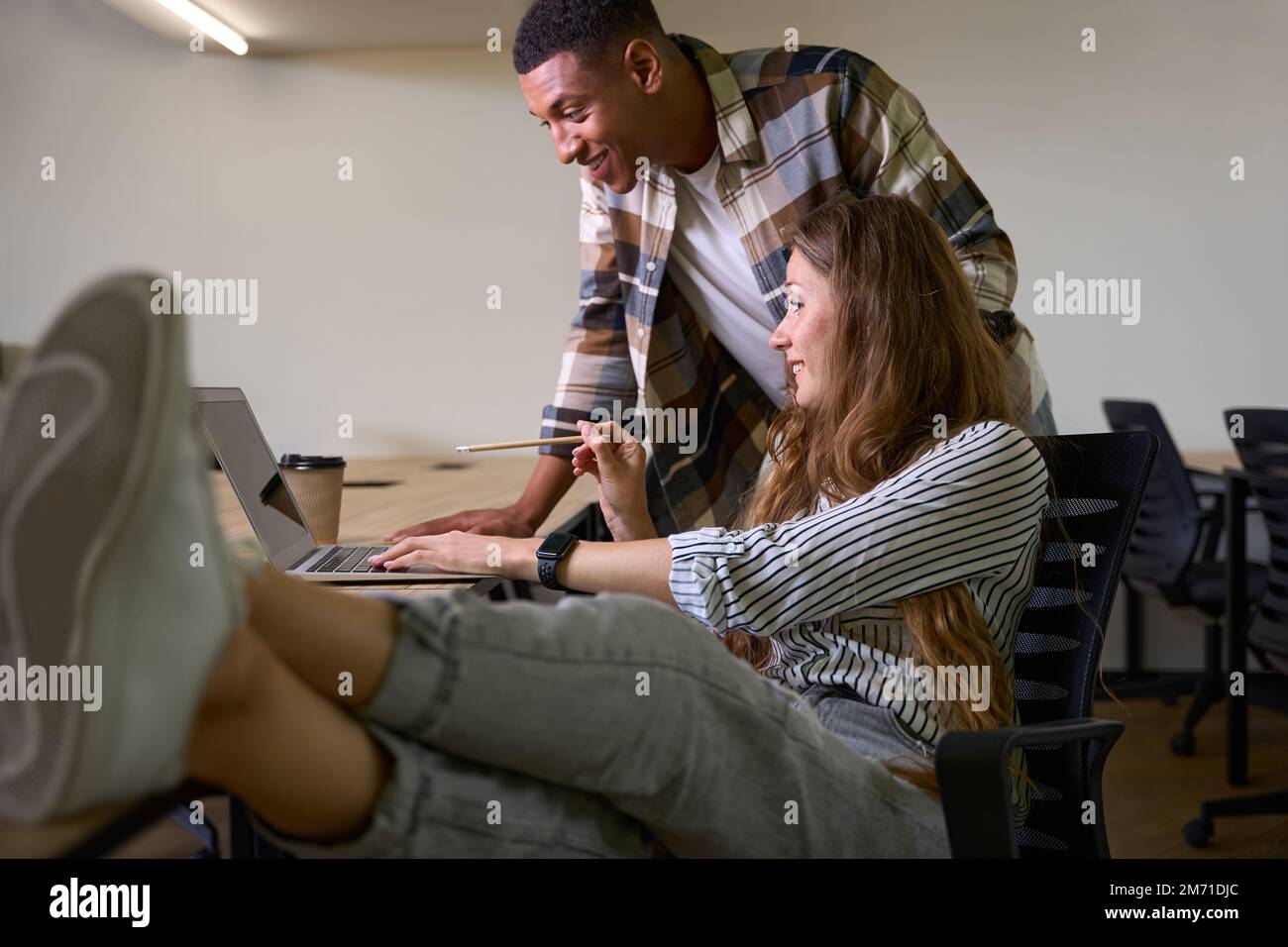 Lächelnde Frau, die Beine auf den Tisch wirft, während sie mit ihrem Kollegen den Laptop benutzt Stockfoto