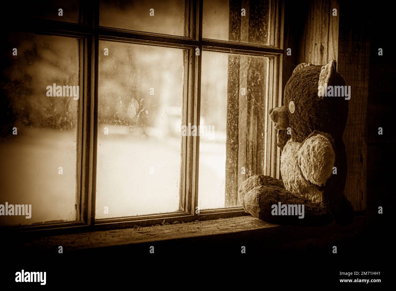 Ein Bild in Sepia-Tönen und Vignettierung eines sehr alten handgefertigten Teddybären, der im Fenster einer verlassenen historischen Pionierhütte sitzt. Stockfoto