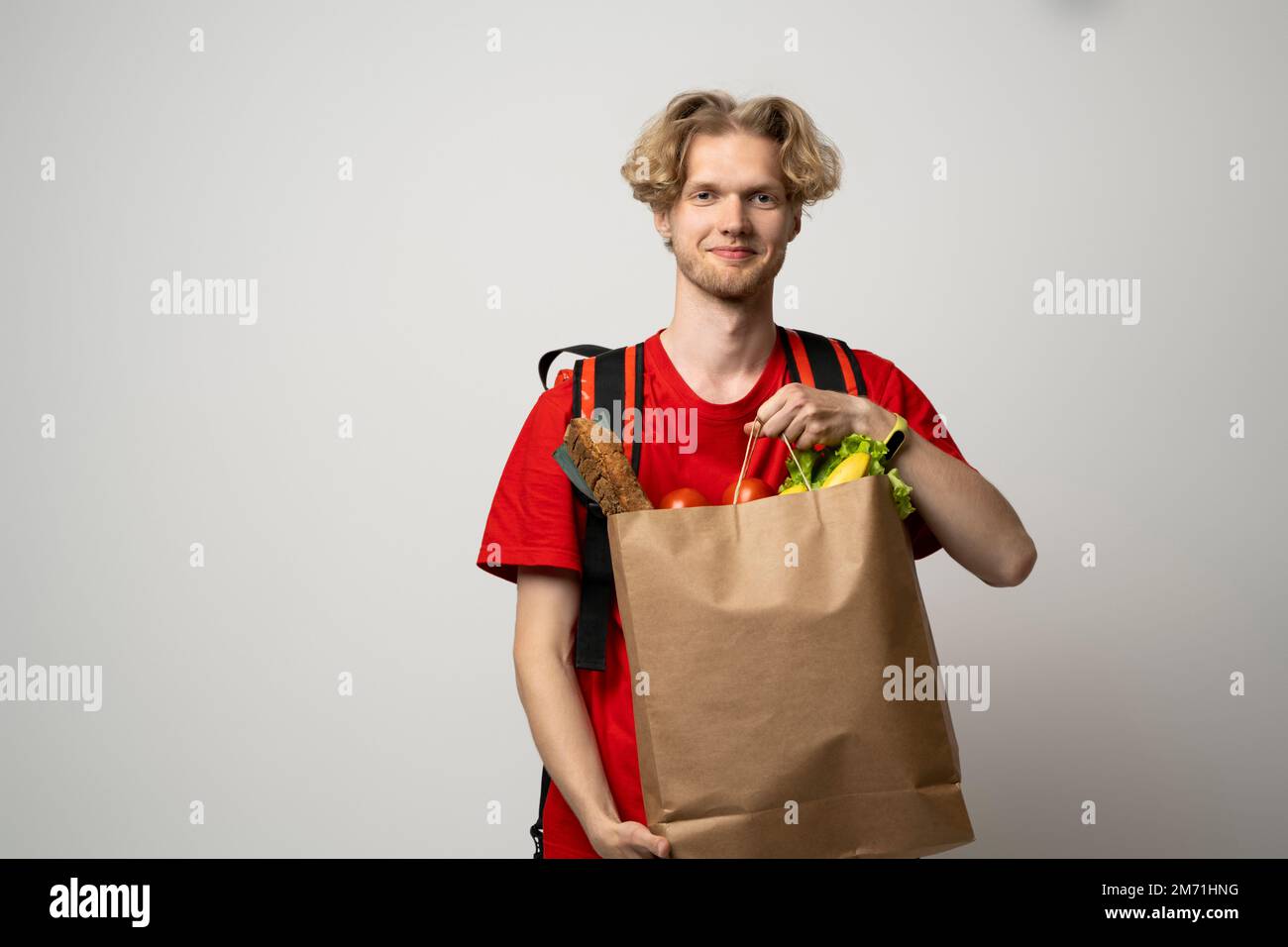 Lieferkonzept. Ein hübscher junger Liefermann in roter Uniform, der eine Packung Lebensmittel und Getränke aus dem Laden trägt, auf dem weißen Studio vermarktet Stockfoto
