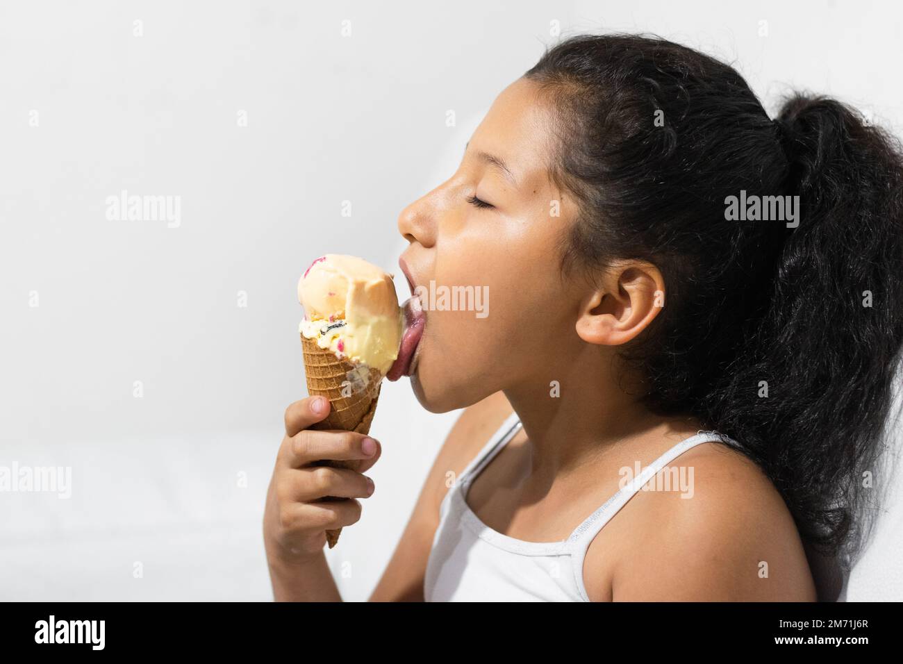 Nahaufnahme eines kleinen lateinischen Mädchens, das ihr Eis leckt, während sie die Augen schließt, und ihr Eis genießt. Mit weißem Hintergrund Stockfoto
