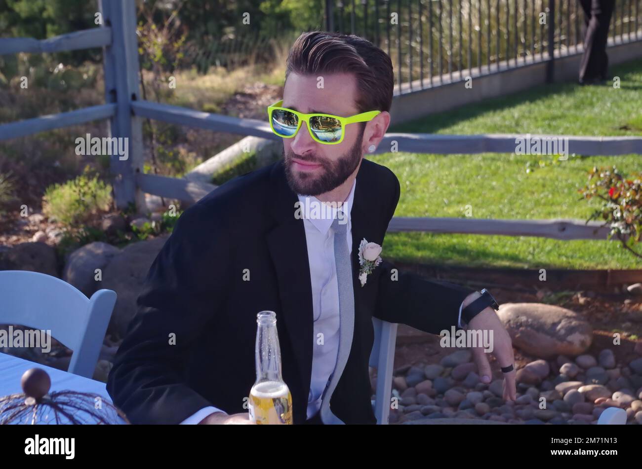 Junger Mann, der an einer feierlichen Veranstaltung teilnimmt, trägt eine bunte Sonnenbrille im Ray-Ban-Stil und trinkt ein Bier Stockfoto