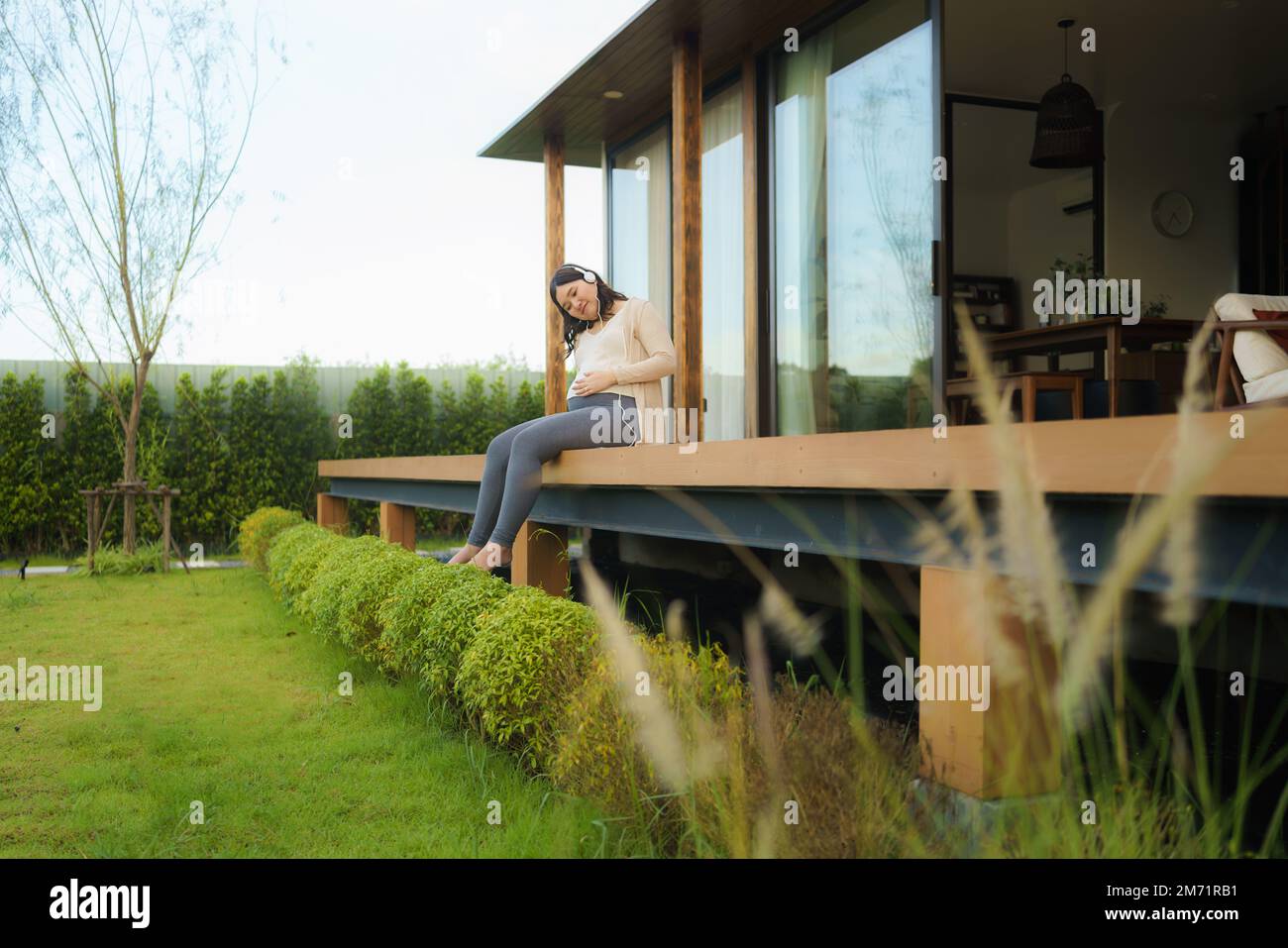 Porträt einer schwangeren Happy Asian Woman, die zu Hause auf der Terrasse sitzt und über ein Smartphone Musik über Kopfhörer hört. Stockfoto