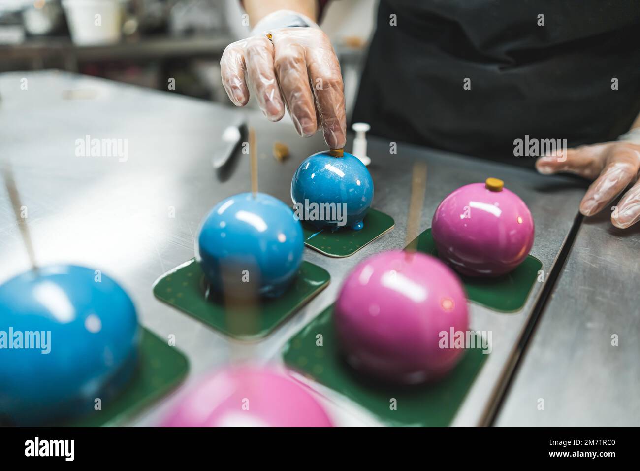 Drei blaue und drei rosa Kuchen auf grünen quadratischen Tellern, die von einem Experten mit transparenten Handschuhen und schwarzer Schürze dekoriert werden. Verschwommener Vordergrund. Hochwertiges Foto Stockfoto
