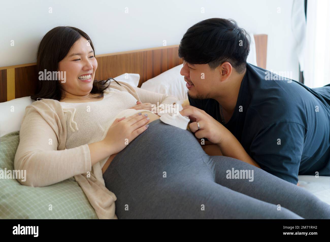 Porträt einer asiatischen, glücklichen schwangeren Frau und ihres Mannes, der Babyschuhe auf dem Bett im Schlafzimmer zu Hause hält Stockfoto