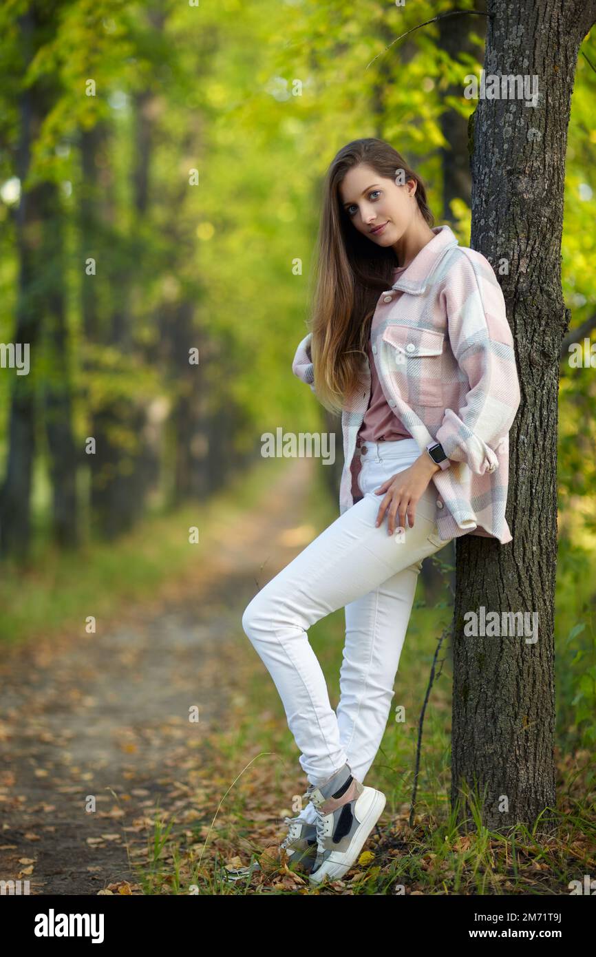 Blonde Frau in blauem Kleid, die sich in einer Herbstlindenallee an einen Baumstamm lehnt. Pfad unter gelben Bäumen mit herbstlichen Blättern. Sonniger Herbst Stockfoto