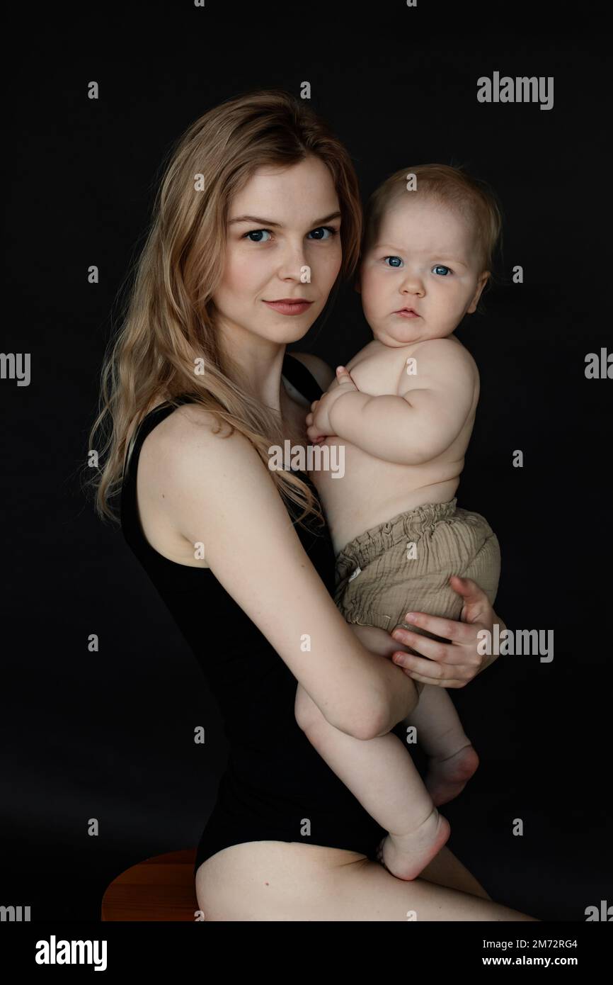 Vertikales Nahaufnahme-Porträt einer jungen blonden Frau, Mutter hält kleines Baby in Windeln in Armen, schau in die Kamera Stockfoto