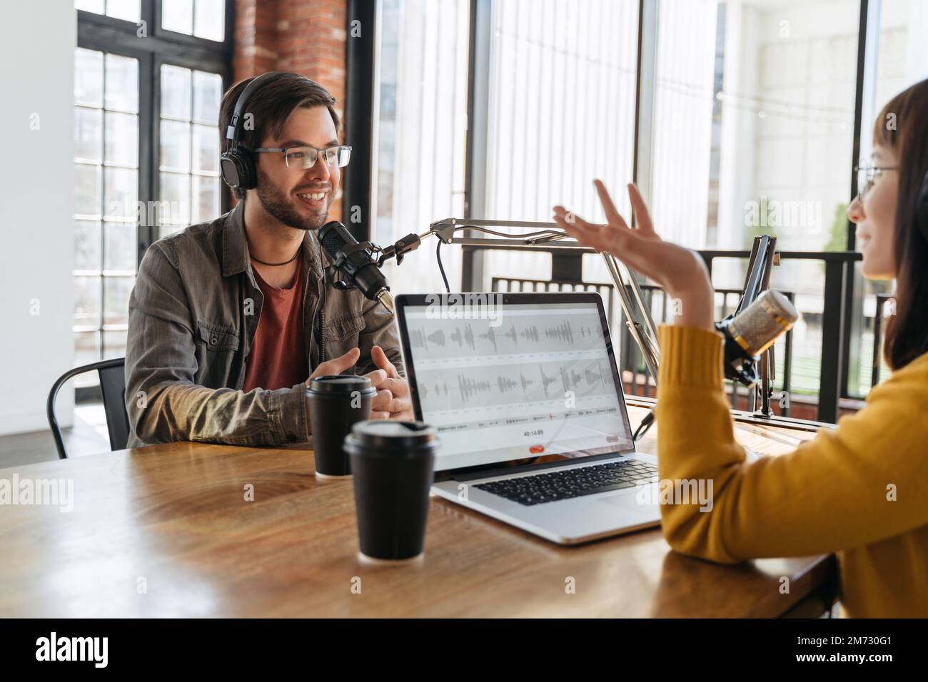 Ein fröhlicher junger Blogger, der mit einer Gesten-Moderatorin spricht, lächelt und ein Interview für einen Audio-Podcast im Studio führt. Junge Männer und Frauen nehmen Livestreams auf. Soziale Medien, Content-Ersteller Stockfoto
