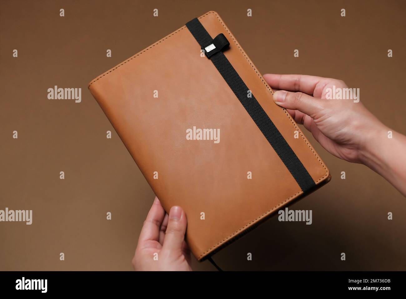 Die Hände halten große braune Lederzeitschrift mit schwarzem Band gegen die braune Oberfläche. Planer für das neue Jahr. Eleganter Organisator. Horizontaler Hintergrund. Stockfoto