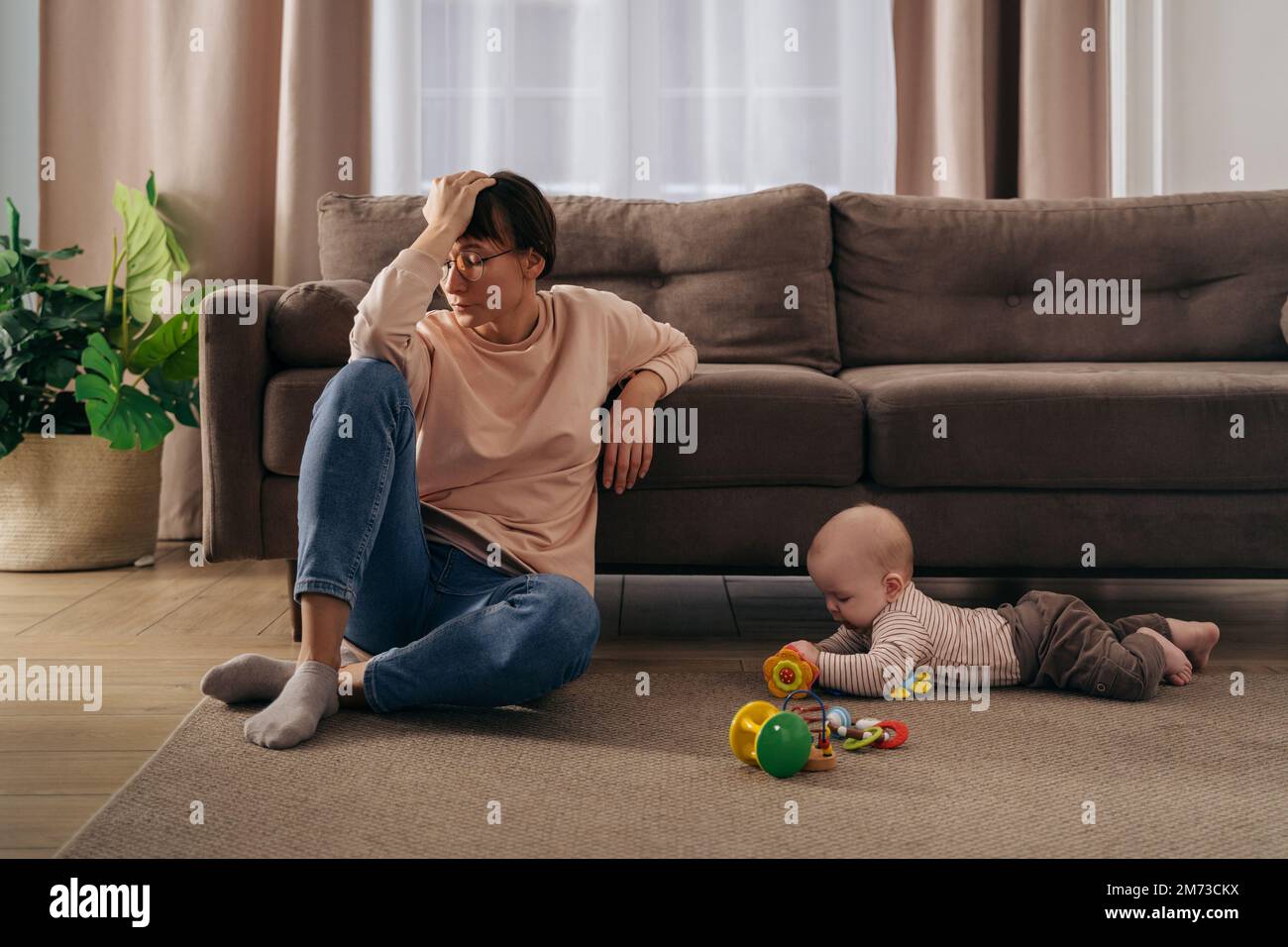 Junge müde Frau, die an postnataler Depression leidet, ihren Kopf mit der Hand hält, auf dem Boden im Wohnzimmer neben ihrem Kleinkinder sitzt und mit Spielzeug spielt. Neue mutter müde von Mutterschaftsschwierigkeiten Stockfoto