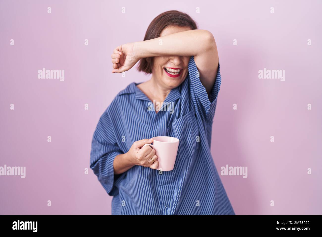 Eine hispanische Frau im mittleren Alter, die eine Tasse Kaffee trinkt, lächelt fröhlich und guckt mit den Händen, die das Gesicht zeigen. Überrascht und verlassen Stockfoto