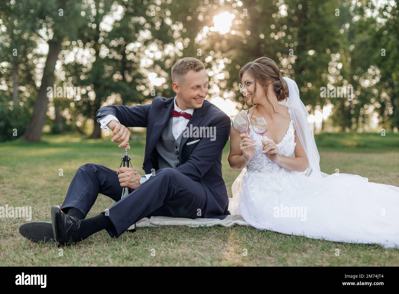 Lächelnde und reizende Bräutigame und Braut mit Brille sehen sich gegenseitig beim Picknick im Park an, offene Weinflasche zum Feiern Stockfoto