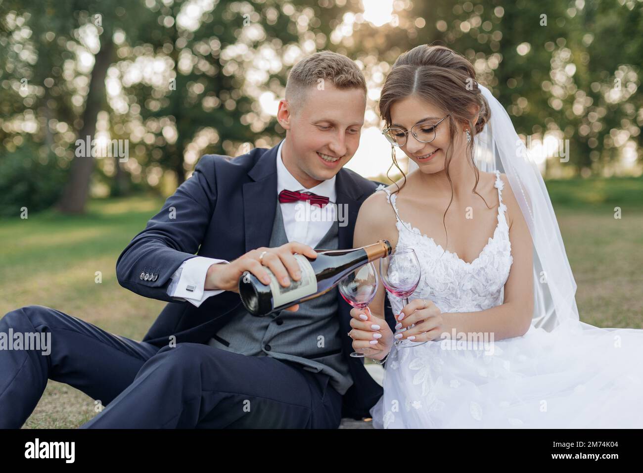 Lächelnder und schüchterner Bräutigam und Braut, trinken und schütten Sekt aus der Flasche in Gläser zusammen, sitzen Sie im Park Stockfoto