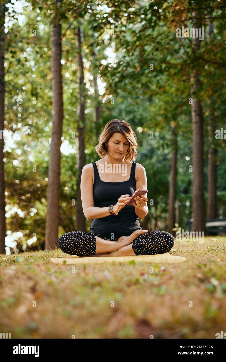 Eine schlanke Jogi-Frau sitzt im Wald in Lotus-Pose und benutzt ein Telefon. Stockfoto