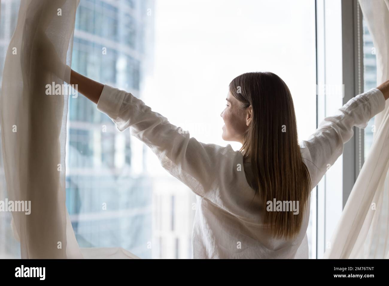 Die Frau öffnet die Vorhänge, begrüßt den neuen Tag, genießt den Blick auf die große Stadt Stockfoto