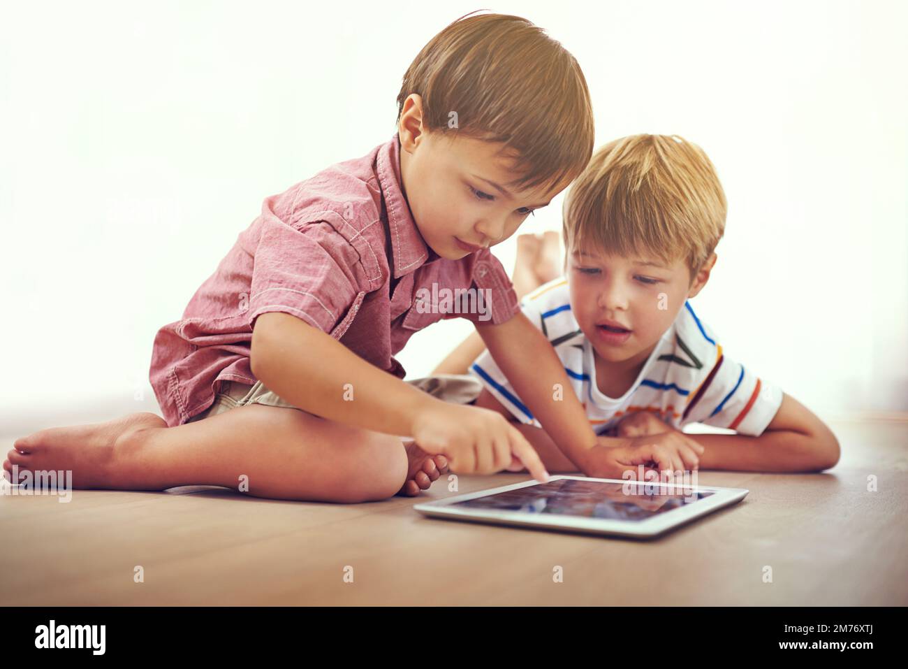 Es gibt so viel, was sie online lernen können. Zwei kleine Jungen, die zusammen ein digitales Tablet benutzen, während sie auf dem Boden sitzen. Stockfoto