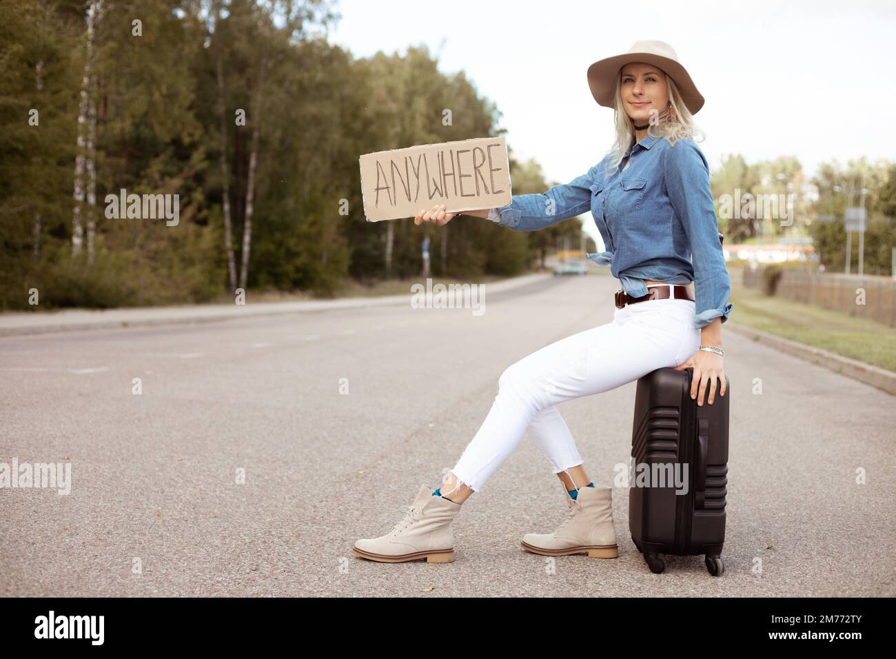Eine nette Frau versucht aufzuhören, vorbeizufahren, indem sie auf einem Koffer mit einem ausgestreckten Pappposter auf einem leeren Highway sitzt. Frau mit Hut, entfliehen Sie der Stadt, um zu gehen Stockfoto