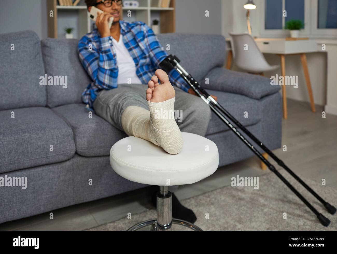 Junger Mann mit verletztem Bein, der zu Hause auf dem Sofa sitzt und telefoniert. Stockfoto