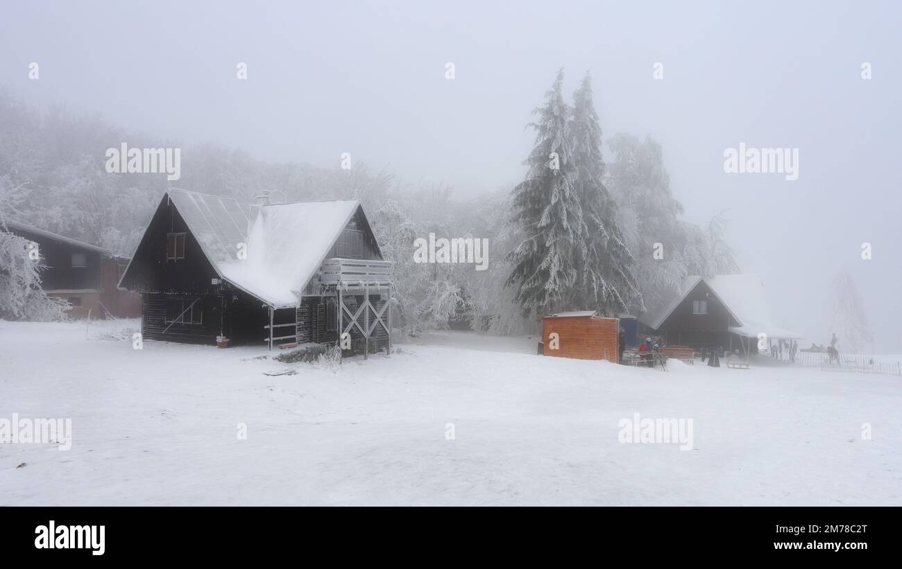 Fantastische Winterlandschaft mit leuchtendem Holzhaus vor der Kulisse der glühenden Lichter der Stadt im Nebel. Gemütliche Hütte in den Karpaten. Weihnachten Stockfoto