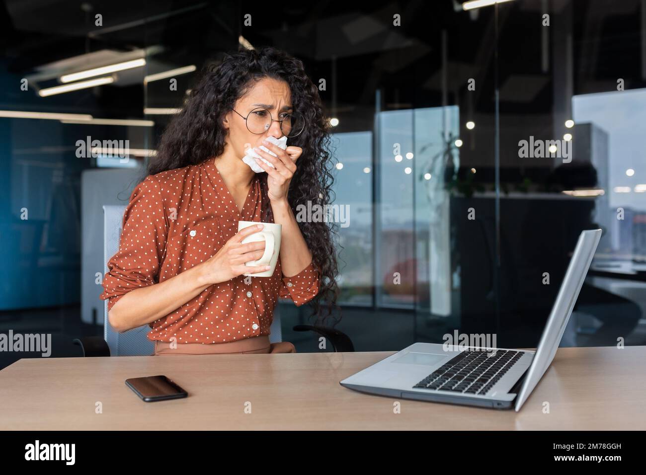 Eine kranke junge Geschäftsfrau aus Lateinamerika, ein Freiberufler, sitzt mit einem Laptop im Büro. Er hält eine Tasse Tee, Medizin in seinen Händen. Er putzt sich die Nase mit einer Serviette, hustet, niest, fühlt sich schlecht an. Stockfoto