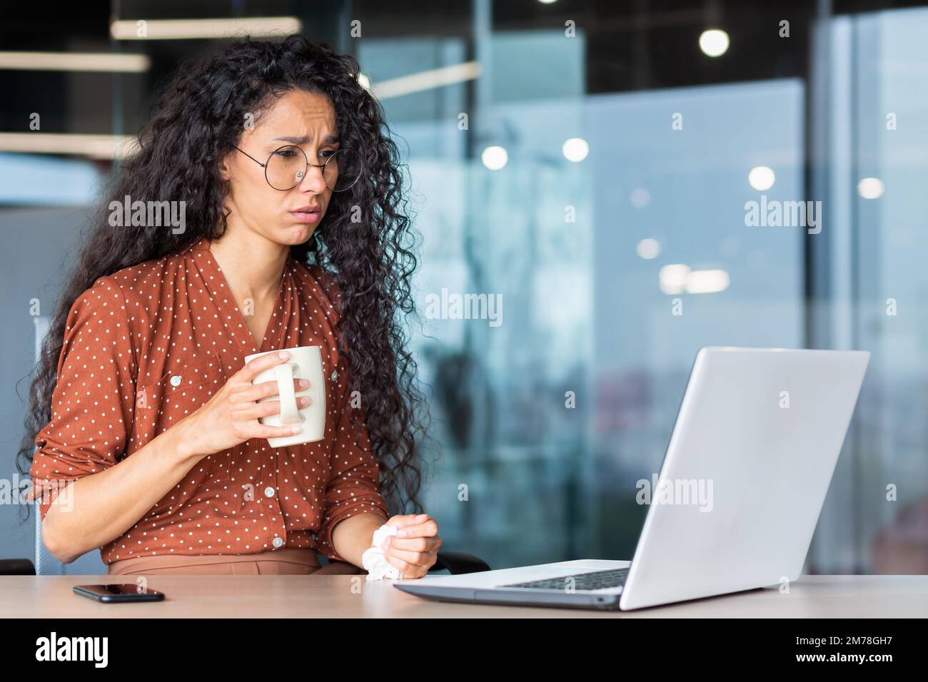 Eine kranke junge Geschäftsfrau aus Lateinamerika, eine Freiberuflerin, sitzt am Schreibtisch im Büro und arbeitet an einem Laptop. Er hält eine Tasse Tee und eine Serviette in den Händen. Sie ist aufgebracht, sie fühlt sich schlecht. Stockfoto