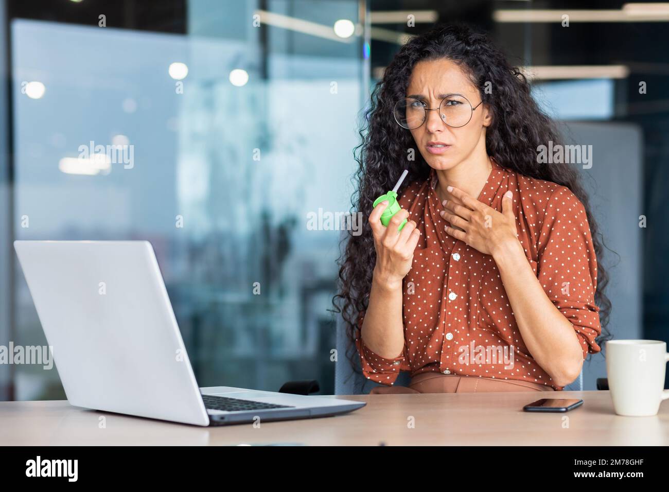 Eine junge kranke lateinamerikanische Frau sitzt im Büro an einem Tisch mit einem Laptop und fühlt sich schlecht. Er hustet, bläst sich die Kehle mit einem Aerosol-Medikament. Er hält seine Brust und schaut traurigerweise in die Kamera. Stockfoto