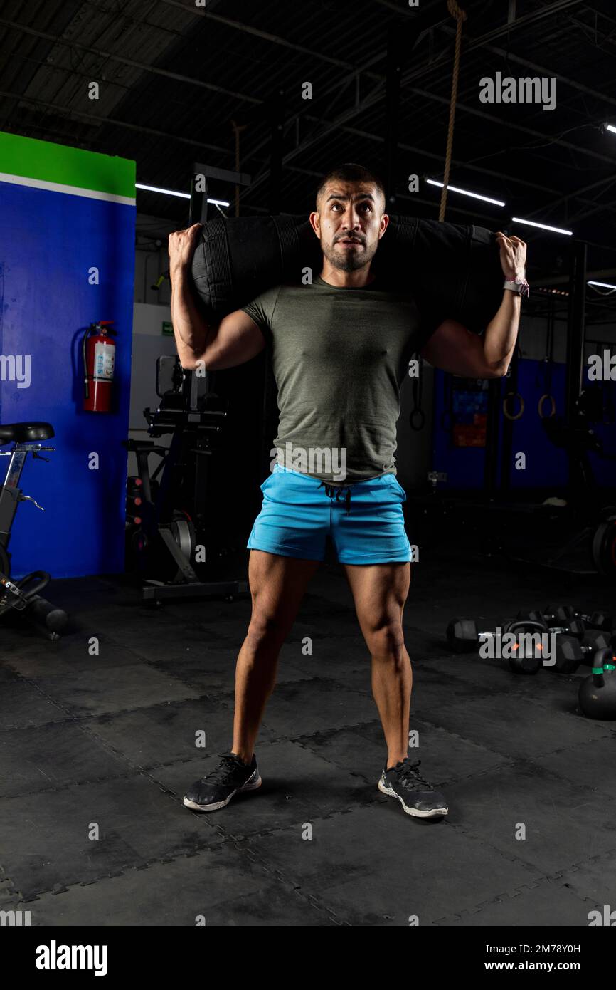Ein Lateiner mit Sportbekleidung, der beim Training im Fitnessstudio einen Sandsack auf dem Rücken trägt Stockfoto